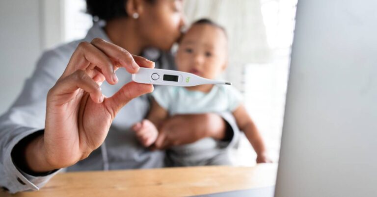 Parent holding a baby while checking a thermometer, deciding when to go to urgent care for a possible fever.